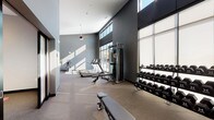 This fitness center is designed to provide a fully functional workout environment within a modern, single-story layout. The space is characterized by soaring vaulted ceilings that lend an open, airy quality throughout, complemented by large floor-to-ceiling windows and elevated clerestory glazing that flood the interior with natural light and offer views of the surrounding area. The walls are finished in a sophisticated palette of charcoal and medium gray tones, paired with durable rubberized flooring suited for high-traffic athletic use.

The facility is divided into distinct functional zones. The cardio area is well-appointed with multiple treadmills, elliptical trainers, upright and recumbent stationary bikes, and rowing machines. A dedicated strength-training section features a multi-station cable machine, a full rack of hex dumbbells, an adjustable weight bench, and additional free-weight accessories. A separate, partitioned room with frosted glass sliding doors provides a more intimate space, finished with light-colored flooring and wall mirrors, suitable for stretching or group exercise. Ceiling fans are installed throughout to maintain airflow, and recessed lighting supplements the generous natural light. The overall ambiance is modern, clean, and purposefully designed for a comprehensive fitness experience.