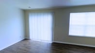 This two-story space offers a clean, functional layout with well-maintained finishes throughout. The living area showcased in the image features rich, dark hardwood-style flooring that contrasts neatly with the light, neutral-toned walls, creating a balanced and inviting atmosphere. A large sliding glass door, dressed with full-length white vertical blinds, serves as a prominent architectural feature, allowing abundant natural light to fill the room while also providing potential access to an outdoor area. A secondary window on the adjacent wall further enhances the brightness of the space. White baseboards frame the perimeter of the room, adding a crisp finishing detail that complements the overall color palette. The ceiling is finished with a subtle texture and includes a visible HVAC vent, indicating a central air system. The open, uncluttered layout of this main living area provides versatility for various furniture arrangements. Spanning two floors, the property offers separation between living and private spaces, a practical design choice that adds functionality and defined zones throughout the home.