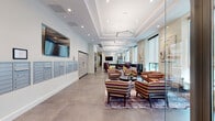 This contemporary residential lobby is thoughtfully designed to serve as both a functional arrival point and a welcoming communal space. The layout extends along a generous corridor, finished with large-format polished concrete tile flooring that adds an understated industrial elegance throughout.

The lounge area is a standout feature, furnished with vibrant multi-striped upholstered armchairs and sofas arranged around a round dark-wood coffee table, all anchored by a patterned area rug. Natural light filters in through large windows fitted with roller shades, while curated abstract artwork adorns the walls, lending the space a refined, gallery-like quality. Recessed lighting and striking geometric pendant fixtures illuminate the tray ceilings, enhancing the sense of height and architectural detail.

Adjacent to the lounge, a sleek reception and concierge desk in dark wood provides a polished focal point, accompanied by a wall-mounted flat-screen TV. The mailroom wall features an extensive bank of individual resident mailboxes in brushed metal, alongside a parcel locker system for added convenience.

A dedicated elevator lobby houses two stainless steel elevators beneath dramatic geometric pendant lighting and soaring ceilings. The space also includes a well-appointed kitchenette with two-tone cabinetry — white upper cabinets paired with wood-grain lower cabinets — and a clean white countertop with a stainless steel sink.