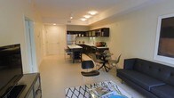 This single-floor space showcases a thoughtfully designed open-concept layout that seamlessly connects the kitchen, dining, and living areas into one cohesive, functional environment. The kitchen is outfitted with dark cabinetry that creates a striking contrast against light-colored walls and flooring, and features stainless steel appliances including a refrigerator and microwave. A kitchen island with bar seating provides additional prep space and casual dining options. Adjacent to the kitchen, the dining area accommodates a contemporary round table with modern molded chairs, making it an ideal spot for everyday meals. The living area is anchored by a streamlined dark sofa paired with a glass coffee table and a geometric-patterned area rug, lending the space a clean, modern aesthetic. Light tile flooring runs throughout, contributing to a bright and airy atmosphere. Recessed lighting and flush-mount ceiling fixtures provide ample, well-distributed illumination across the entire space. Track lighting near the entryway adds a practical and stylish element. The front entry door is clearly defined, offering a welcoming transition into the home. Overall, the space presents a modern, urban sensibility with a neutral palette and contemporary furnishings that balance style and practicality.