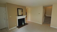 This two-floor home offers a thoughtfully arranged layout that balances comfort and functionality. The main living area serves as the heart of the space, anchored by a classic wood-mantled fireplace with a dark surround and a tiled hearth, adding warmth and character to the room. A decorative mirror mounted above the mantle enhances the sense of space, while the neutral beige and cream wall tones create a calm, welcoming atmosphere throughout.

Soft carpeting covers the main living area, transitioning to tile flooring near the fireplace hearth and adjoining spaces. The open sightline from the living area leads directly into the kitchen, which is visible in the background and features light cabinetry and tile flooring, suggesting a practical and connected layout ideal for everyday living.

A staircase is visible along one wall, providing access to the second floor and hinting at additional living space above. Closet access near the staircase adds convenient storage to the main level. The overall design is clean and understated, with simple white trim and baseboards framing each room. Natural light filters into the space, contributing to the bright, airy feel of the interior. The home's neutral palette and traditional architectural details make it a versatile and comfortable residence.