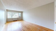 This single-floor unit is thoughtfully laid out to maximize its compact footprint, comprising a living room, kitchen, bathroom, and hallway. The living room is the standout space, featuring warm-toned hardwood flooring that runs throughout and a textured popcorn ceiling that adds a classic character. A large window with an in-unit air conditioning unit allows natural light to fill the room while offering views of the surrounding greenery outdoors. A radiator beneath the window provides supplemental heating. The neutral white walls offer a clean, versatile backdrop throughout the space. The kitchen is equipped with white cabinetry, a double stainless steel sink, richly patterned stone-look countertops, and a white range, all complemented by tile flooring and a window fitted with horizontal blinds that brings in additional natural light. The bathroom features tile-surround walls around the tub and shower, tile flooring, and clean white fixtures. A hallway connects the rooms efficiently, with a warm-toned wood front entry door adding a touch of character at the entrance. Overall, the unit presents a straightforward and functional layout well-suited for everyday living.
