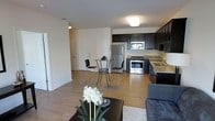 This single-floor residence offers a thoughtfully designed open-concept layout that unifies the living, dining, and kitchen areas into one cohesive and functional space. The interior is finished with warm hardwood-style flooring that extends throughout, lending a sense of continuity and warmth to the entire floor plan.

The kitchen is well-appointed with dark espresso cabinetry that provides ample storage, light granite countertops, a stainless steel refrigerator, and a stainless steel range with oven — all contributing to a sleek, modern aesthetic. A double sink sits beneath a window, allowing natural light to brighten the workspace.

Adjacent to the kitchen, the dining area comfortably accommodates a bistro-style table and chairs, making it an ideal spot for casual meals. The living area features plush seating and a cozy area rug, creating an inviting space to relax and unwind.

The neutral color palette of white walls and dark furnishing accents strikes a clean, contemporary balance throughout. A hallway leads to additional rooms, offering separation and privacy from the main living spaces. Overall, this residence combines modern finishes with a practical, open layout that maximizes comfort and livability.