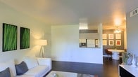 This single-floor space offers an open and functional layout defined by dark hardwood flooring and clean, neutral-toned walls. The living area is spacious and bright, illuminated by a stylish tripod floor lamp that adds a warm, ambient glow. A white sofa anchors the space, contributing to the minimalist aesthetic. A defining architectural feature is the half-wall partition with a pass-through window that connects the living area to the kitchen, allowing light and sightlines to flow between the two spaces while maintaining a degree of separation. The kitchen, visible through the pass-through, features white cabinetry and a white refrigerator, consistent with the clean, light color palette throughout. Fluorescent lighting in the kitchen provides functional illumination. A doorway off the living area leads to additional rooms, and a second adjoining area is visible to the right, suggesting a versatile, connected floor plan. The overall ambiance is clean, understated, and practical, with a layout that maximizes openness across the single floor.