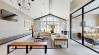 This thoughtfully designed single-story space blends contemporary aesthetics with warm, inviting elements to create a refined and functional environment. The architecture is immediately impressive, highlighted by dramatic vaulted ceilings with exposed wood beams that anchor the open layout while lending organic warmth to the otherwise crisp, modern interior.

Large-format polished tile flooring flows continuously throughout, reflecting natural light and reinforcing the sense of openness. Floor-to-ceiling steel-framed windows and glass walls allow generous views of the surrounding greenery, flooding each area with daylight. A prominent arched window serves as a striking focal point in the main lounge, framing the outdoor landscape beautifully.

The lounge area features plush neutral sofas, a linear fireplace, curated wall art displayed on floating shelves, and an eclectic mix of sculptural pendant lighting. Adjacent, a dedicated workspace offers a substantial communal table with modern seating, built-in shelving, and workstations backed by an artisan mixed-media wall display.

A secondary sitting area showcases a geometric gold-accented credenza, glass coffee table, and tall windows overlooking a landscaped exterior terrace. Throughout, the carefully layered lighting design—ranging from clustered pendants to wall sconces—enhances the space's contemporary yet welcoming ambiance.