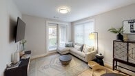 This single-floor residence is thoughtfully designed with a neutral palette of soft grays and whites, complemented throughout by light wood-tone flooring that lends warmth and continuity to each space. The living area is anchored by a corner sectional sofa and benefits from generous natural light streaming through large windows and a glass-paneled exterior door, offering views of the surrounding greenery. The kitchen is a functional highlight, featuring two-tone cabinetry—white upper cabinets paired with slate-gray lower cabinets—white subway tile backsplash, light quartz countertops, stainless steel appliances including a gas range, dishwasher, and side-by-side refrigerator, and open shelving for additional storage. The bedrooms are comfortably sized; the primary bedroom features a ceiling fan and a large window with sheer curtains that invite soft, diffused light, while the secondary bedroom includes a ceiling fan and direct door access. The full bathroom is finished with large-format gray tile surrounding a tub-shower combination, a framed mirror, and white cabinetry with gray quartz countertops. A dedicated home office features wall-mounted open shelving on either side of a central workstation, with a large-scale landscape painting adding an artistic focal point to the room.
