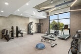This fitness center offers a thoughtfully organized workout space across a single floor, designed to accommodate a diverse range of exercise needs. The space combines modern finishes with industrial architectural elements, including exposed concrete block walls and visible utility infrastructure, creating a functional and contemporary aesthetic throughout.

The flooring consists of durable speckle-finish rubber or epoxy material suited for high-traffic gym use. A large floor-to-ceiling window along one wall floods the cardio zone with natural light while providing street-level views, lending an open, energizing atmosphere to the space. Linear LED lighting fixtures run across the ceiling, supplemented by recessed downlights, ensuring consistent, even illumination throughout.

Equipment is well-distributed across distinct zones. The cardio area houses a treadmill, elliptical trainer, stationary spin bike, and a rowing machine. The strength zone features selectorized weight machines, including leg extension and multi-functional cable units. The free-weight area includes a full tiered dumbbell rack, a kettlebell rack with multiple weights, a flat utility bench, a BOSU balance trainer, and foam roller accessories. A mounted flat-screen TV enhances the workout environment. Two interior doors are accessible along one wall, and a fire panel is visibly installed, indicating proper safety compliance.