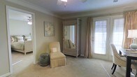 This single-floor space exudes a calm, refined ambiance through its cohesive neutral color palette and thoughtful design elements. The room showcased features crown molding that frames the ceiling, adding a touch of classic elegance to the space. Soft, wall-to-wall carpeting in a warm beige tone creates a comfortable and inviting atmosphere underfoot.

A standout piece in the room is a distressed, ornate armoire with a full-length mirror, serving as both a functional storage unit and a decorative focal point. Beside it, a cushioned chaise lounge chair in a complementary cream tone offers a cozy seating area, accompanied by a ceramic garden stool that doubles as a side table. Floral artwork on the wall reinforces the soft, neutral aesthetic.

Natural light enters generously through a large window dressed with floor-length linen drapes, which also frames a glass door offering outdoor access. The overall ambiance is tranquil and sophisticated, with layered textures and subtle décor creating a cohesive, relaxing environment throughout the space.