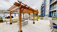 Located at 101 Viva Bene Circle in Saint Peters, Missouri, this outdoor patio space serves as a well-appointed communal gathering area adjacent to a modern multi-story residential building. The patio features a spacious concrete surface that accommodates multiple distinct seating zones, providing ample room for both social gatherings and relaxed outdoor living.

At the heart of the space stands a substantial natural wood pergola, constructed with robust timber beams and decorative joinery. Beneath it, a stone-surround fire pit anchors a lounge area furnished with cushioned wicker sofas and chairs, creating an inviting setting for conversation and relaxation. The pergola's open-lattice roof allows filtered light while offering a sense of shelter.

Throughout the patio, multiple dining tables are arranged with coordinating wood-and-metal chairs, each set featuring navy blue cushions that echo the bold blue tones of the adjacent building's exterior. Large navy market umbrellas provide shade over several of the dining groupings, enhancing comfort during outdoor use.

The surrounding landscape includes neatly maintained planting beds with young shrubs and mulched borders, contributing a clean, finished aesthetic to the space. The patio connects directly to the building's ground-floor entrance, with balconies visible overhead on the building façade. The overall design balances functional outdoor dining with relaxed lounge amenities, making it a versatile and well-equipped exterior amenity space.