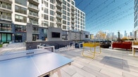Located at 5 Port Imperial Blvd in Weehawken, NJ, this outdoor amenity deck offers a thoughtfully designed resort-style experience with sweeping views of the New York City skyline. The centerpiece of the space is a generously sized swimming pool finished with elegant blue mosaic tile, surrounded by a broad sun terrace lined with lounge chairs and bold black-and-white striped umbrellas, creating a vibrant, resort-inspired atmosphere.

The deck is organized into distinct activity and relaxation zones. A dedicated game area features a ping-pong table, oversized chess set, and foosball table, all set beneath festoon string lighting that adds ambiance during evening hours. Mounted flat-screen TVs and an outdoor bar with counter seating make this area ideal for social gatherings.

Multiple lounge seating arrangements are distributed throughout the deck, including plush blue sofas, modular armchairs, and bistro tables with yellow accents that inject a playful pop of color. A covered terrace area with floor-to-ceiling glass walls connects seamlessly to an interior amenity lounge, with large sliding glass doors that blur the boundary between indoors and outdoors.

The surrounding architecture features a contemporary mix of brick and glass tower facades, complementing the polished stone pavers underfoot. Lush planters and greenery are integrated throughout, softening the urban environment and enhancing the overall sense of retreat.