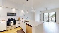 Located at 8205 Commerce Way in Miami Lakes, FL, this modern 2-bedroom, 2-bathroom residence offers a thoughtfully designed open-concept layout with clean, contemporary finishes throughout. Light-toned wood-style flooring runs seamlessly across the entire space, creating a warm and cohesive aesthetic.

The kitchen serves as the centerpiece of the main living area, featuring sleek white flat-front cabinetry, a white quartz waterfall island with an undermount sink, and a full suite of stainless steel appliances including a French door refrigerator, range, microwave, and dishwasher. A trio of globe pendant lights hang above the island, adding a stylish focal point. White subway tile backsplash and tall upper cabinets with open shelving complete the modern look. The kitchen flows directly into the living area, which benefits from high ceilings and access to an outdoor balcony, bringing in abundant natural light.

Both bedrooms are bright and well-proportioned, with large black-framed windows offering views of swaying palm trees and the surrounding South Florida landscape. Each bedroom is served by a dedicated closet with built-in wire shelving. The bathrooms are finished in a clean, all-white palette, with one featuring a soaking tub and shower combination with shiplap-style tile surround and a white vanity with quartz countertop.
