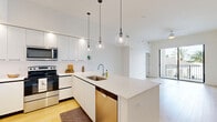 Located at 8205 Commerce Way in Miami Lakes, FL, this modern 2-bedroom, 2-bathroom residence offers a thoughtfully designed open-concept layout with clean, contemporary finishes throughout. Light-toned wood-style flooring runs seamlessly across the entire space, creating a warm and cohesive aesthetic.

The kitchen serves as the centerpiece of the main living area, featuring sleek white flat-front cabinetry, a white quartz waterfall island with an undermount sink, and a full suite of stainless steel appliances including a French door refrigerator, range, microwave, and dishwasher. A trio of globe pendant lights hang above the island, adding a stylish focal point. White subway tile backsplash and tall upper cabinets with open shelving complete the modern look. The kitchen flows directly into the living area, which benefits from high ceilings and access to an outdoor balcony, bringing in abundant natural light.

Both bedrooms are bright and well-proportioned, with large black-framed windows offering views of swaying palm trees and the surrounding South Florida landscape. Each bedroom is served by a dedicated closet with built-in wire shelving. The bathrooms are finished in a clean, all-white palette, with one featuring a soaking tub and shower combination with shiplap-style tile surround and a white vanity with quartz countertop.