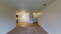 This single-floor space presents an open-concept layout that connects the living, dining, and kitchen areas in a cohesive and functional arrangement. The living area is finished with neutral-toned carpet, lending a comfortable and understated feel to the space. A textured ceiling runs throughout, adding a subtle architectural detail to the otherwise clean, white walls.

The dining area is defined by a classic chandelier that serves as a focal point, providing warm ambient lighting. A partial wall separates the dining space from the adjacent kitchen, which features warm-toned wood cabinetry and tile flooring that transitions naturally from the carpeted areas. The kitchen area is compact yet organized, with upper and lower cabinetry providing practical storage.

A hallway visible at the rear of the space leads to additional rooms, with a door hinting at further private areas beyond. Ceiling vents are positioned throughout, indicating a central HVAC system. The overall ambiance is neutral and understated, offering a functional and move-in-ready environment well-suited to a variety of furnishing styles.