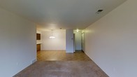 This single-floor space presents an open-concept layout that connects the living, dining, and kitchen areas in a cohesive and functional arrangement. The living area is finished with neutral-toned carpet, lending a comfortable and understated feel to the space. A textured ceiling runs throughout, adding a subtle architectural detail to the otherwise clean, white walls.

The dining area is defined by a classic chandelier that serves as a focal point, providing warm ambient lighting. A partial wall separates the dining space from the adjacent kitchen, which features warm-toned wood cabinetry and tile flooring that transitions naturally from the carpeted areas. The kitchen area is compact yet organized, with upper and lower cabinetry providing practical storage.

A hallway visible at the rear of the space leads to additional rooms, with a door hinting at further private areas beyond. Ceiling vents are positioned throughout, indicating a central HVAC system. The overall ambiance is neutral and understated, offering a functional and move-in-ready environment well-suited to a variety of furnishing styles.