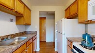 Located at 681 Babbitt Rd in Euclid, Ohio, this two-bedroom, one-bathroom unit offers a practical and well-organized single-floor layout. The living area features hardwood-style flooring and multiple windows that invite natural light and outdoor views of the surrounding neighborhood. A galley-style kitchen runs along both walls, lined with warm honey-toned wood cabinetry, dark granite-look countertops, a stainless sink, and white appliances including a full-size refrigerator and range. The kitchen connects seamlessly to the rest of the unit via a central hallway. Both bedrooms are carpeted and feature windows with blinds, providing a comfortable, quiet retreat. The primary bedroom is a generous size with ample wall space. A carpeted hallway with louvered interior doors connects the bedrooms to the full bathroom, which is a standout feature with its retro turquoise tile work covering the walls and mosaic-tiled floor, a soaking tub with shower, and a white vanity. Throughout the unit, neutral wall tones create a clean, cohesive aesthetic. Two hallways provide efficient circulation throughout the space, making the layout feel functional and easy to navigate.