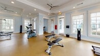 This fitness center in Pooler, Georgia offers a well-appointed, single-floor workout facility designed with both function and aesthetics in mind. The space is characterized by soft powder-blue walls, crisp white crown molding, and tray ceilings accented with recessed lighting and sleek ceiling fans — creating an inviting, airy atmosphere that sets it apart from a typical gym environment.

Large, white-trimmed windows are positioned throughout the space, flooding the interior with abundant natural light and providing views of the surrounding greenery. The durable speckled flooring runs throughout, offering both practicality and a clean, cohesive look underfoot.

The facility is comprehensively equipped for a full-body workout. Cardio equipment includes Matrix treadmills and elliptical machines, while the strength training area features a Hoist cable machine, a multi-function weight station, adjustable benches, and a medicine ball rack. Wall-mounted flat-screen televisions provide entertainment during workouts.

A central open area allows for flexible training and free movement between equipment zones. Additional amenities include restroom access, an exit door with outdoor access, and a sanitization station. The thoughtful layout ensures ample space between equipment, allowing for a comfortable and safe exercise experience throughout the facility.