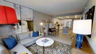 This single-floor residence offers a well-designed open-concept layout that effortlessly connects the living, dining, and kitchen areas into one cohesive and functional space. The living area is anchored by a contemporary light grey sofa, a round marble-top coffee table, and a bold multicolored chevron area rug that adds a lively focal point to the room. A large-scale abstract artwork adorns one wall, contributing to the space's modern aesthetic.

The dining area sits centrally within the open floor plan, featuring a compact dining table with seating for several guests, positioned adjacent to a white shelving unit that provides both storage and display space. Natural light flows throughout, complemented by recessed ceiling lighting that ensures the entire area remains bright and welcoming.

The kitchen is thoughtfully designed with white shaker-style cabinetry, a grey island that doubles as additional prep and seating space, stainless steel appliances including a refrigerator and microwave, and light-toned countertops. The kitchen's clean lines and neutral palette create a crisp, modern feel.

Light-toned hardwood-style flooring runs continuously throughout the space, unifying each zone and enhancing the overall sense of openness. The combination of neutral walls, vibrant accents, and modern fixtures gives this residence a balanced, inviting, and contemporary ambiance.