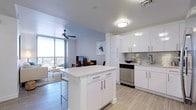 This contemporary single-floor residence is designed with a clean, modern aesthetic throughout, defined by a consistent neutral palette of whites and soft grays. The open-concept kitchen serves as a central hub, featuring high-gloss white cabinetry, light quartz countertops, a metallic subway tile backsplash, and a full suite of stainless steel appliances including a range, microwave, dishwasher, and French door refrigerator. A central island with bar seating adds both workspace and a casual dining option. Light wood-look tile flooring extends throughout, tying the living spaces together seamlessly. The living area is bright and airy, anchored by floor-to-ceiling sliding glass doors that open onto a private balcony, flooding the room with natural light and offering outdoor views. A ceiling fan enhances comfort in both the living room and bedroom. The bedroom is calm and well-lit, with large windows dressed in elegant draping curtains. The bathrooms are finished in an equally clean style, featuring white subway tile surrounds, quartz vanity tops, white cabinetry with sleek hardware, and large mirrors. The overall ambiance of the space is modern, light-filled, and thoughtfully appointed, creating a polished and comfortable living environment.