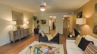 This single-floor residence features an inviting open-concept layout that connects the living area to the kitchen, creating a seamless flow throughout the space. The living room is anchored by warm hardwood flooring and highlighted by a ceiling fan that adds both function and character overhead. A striking exposed brick wall on one side introduces a rustic, textural contrast to the otherwise smooth, neutral-toned walls, lending the space a distinctive architectural charm.

The living area is generously proportioned, comfortably accommodating a dark upholstered sofa set arranged around a round glass coffee table atop a patterned area rug. Soft, ambient lighting from a table lamp on a mirrored sideboard adds warmth to the room. A tall indoor palm-like plant introduces a natural, organic element to the corner of the space.

A pass-through opening connects the living area to the kitchen, which features warm wood cabinetry, a white refrigerator, and light countertops. The hallway leading toward the back of the unit reveals additional rooms bathed in warm lighting. Overall, the space presents a cozy, well-appointed atmosphere with a blend of rustic and contemporary design elements.