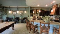 This single-floor space offers a thoughtfully designed open-concept layout that seamlessly blends functional areas with sophisticated interior design. The space is anchored by a generous granite-topped bar counter, surrounded by ornate wooden bar-height chairs that invite gathering and conversation. Pendant lights with wrought-iron fixtures hang overhead, casting warm, ambient light throughout the area.

Adjacent to the bar, a fully equipped kitchen features dark espresso cabinetry, open shelving displaying decorative accents and greenery, and a classic tile backsplash with intricate detailing. The kitchen is well-appointed with modern appliances and ample counter space, making it ideal for both casual use and entertaining.

A dedicated workstation area is positioned nearby, outfitted with built-in desks, computer stations, and task chairs — providing a productive environment within the same open footprint. Framed artwork adorns the soft teal-hued walls, adding a refined, curated feel to the space.

The flooring features a patterned tile with an ornate motif that ties the design elements together cohesively. The overall ambiance is warm and elegant, blending dark wood tones with soft blue-green walls and layered lighting to create an inviting and versatile environment.