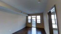 This single-floor space presents a clean, open layout with a neutral color palette that creates a bright and welcoming atmosphere. The main living area is characterized by smooth white ceilings and warm-toned walls that complement the rich, dark hardwood flooring, which adds a sense of warmth and sophistication to the space.

Natural light is a standout feature, streaming in through a pair of windows flanking a central glass door that leads directly to a private balcony. The balcony access provides outdoor views of the surrounding building exteriors, offering a functional extension of the living area. The windows ensure the interior remains well-lit throughout the day.

Ceiling-mounted light fixtures and a visible HVAC vent indicate thoughtful utility integration, maintaining a streamlined aesthetic without compromising functionality. The walls are largely unadorned, offering a versatile canvas for personalization. Baseboard trim runs along the perimeter of the room, adding a subtle architectural detail that frames the space neatly.

Overall, the space conveys a minimalist, move-in-ready character, well-suited for a variety of furnishing arrangements and interior styles.