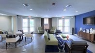 This single-floor space presents a thoughtfully designed communal lounge area that combines comfort and style in an open, airy layout. The room is bathed in natural light streaming through multiple large windows and glass-paneled doors, which also offer views of the surrounding outdoor greenery. Recessed ceiling lighting complements the natural light, ensuring the space remains bright and inviting at all times.

The interior is furnished with a contemporary aesthetic, featuring a mix of seating arrangements including a chartreuse sofa accented with decorative pillows, paired wingback chairs in complementary neutral tones, and additional accent chairs and side seating arranged throughout. A bold navy and yellow geometric area rug anchors the central seating area, adding a vibrant focal point to the polished concrete flooring beneath.

Draped curtains in warm brown tones frame the windows and doors, adding texture and warmth to the otherwise modern palette. Decorative accessories, including sculptural accents and table displays, contribute to the space's curated yet approachable character. The open floor plan allows for flexible use of the space, making it well-suited for casual gatherings, socializing, or relaxed individual use. Overall, the space strikes a balance between functional design and welcoming ambiance.