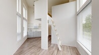 Located at 2211 SW Fourth Ave in Portland, Oregon, this efficiently designed loft-style unit makes striking use of vertical space through dramatically high ceilings that amplify the sense of openness well beyond what the footprint alone would suggest. The main living area is bathed in natural light courtesy of floor-to-ceiling windows fitted with blinds, offering direct views of the surrounding Portland cityscape and greenery. A sleek loft ladder leads to an elevated sleeping or storage area overhead, adding a distinctive architectural element while maximizing the use of vertical space. The kitchenette is neatly appointed with gray cabinetry, open shelving, a sink, and a microwave, providing essential functionality in a compact layout. Light wood-tone vinyl plank flooring runs throughout the main living area, contributing a warm, contemporary aesthetic that complements the crisp white walls. An exterior glass door opens to additional natural light and urban views. A dedicated hallway connects to the bathroom, which features wood-tone flooring, clean white walls, a toilet, and a towel bar. The overall design reflects a modern, minimalist sensibility, making efficient and thoughtful use of every square foot in an urban Portland setting.