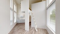 Located at 2211 SW Fourth Ave in Portland, Oregon, this efficiently designed loft-style unit makes striking use of vertical space through dramatically high ceilings that amplify the sense of openness well beyond what the footprint alone would suggest. The main living area is bathed in natural light courtesy of floor-to-ceiling windows fitted with blinds, offering direct views of the surrounding Portland cityscape and greenery. A sleek loft ladder leads to an elevated sleeping or storage area overhead, adding a distinctive architectural element while maximizing the use of vertical space. The kitchenette is neatly appointed with gray cabinetry, open shelving, a sink, and a microwave, providing essential functionality in a compact layout. Light wood-tone vinyl plank flooring runs throughout the main living area, contributing a warm, contemporary aesthetic that complements the crisp white walls. An exterior glass door opens to additional natural light and urban views. A dedicated hallway connects to the bathroom, which features wood-tone flooring, clean white walls, a toilet, and a towel bar. The overall design reflects a modern, minimalist sensibility, making efficient and thoughtful use of every square foot in an urban Portland setting.