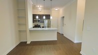 This single-floor residence offers a thoughtfully designed open-concept layout that seamlessly connects the kitchen and living areas. The kitchen is a standout feature, boasting rich dark cabinetry that contrasts beautifully with light granite countertops. Stainless steel appliances, including a refrigerator, microwave, and range, lend a sleek, contemporary feel. A peninsula counter with pendant lighting overhead serves as both a functional workspace and a casual dining or gathering spot, with stylish hanging lights adding warmth and character. The floors throughout are finished in a rich hardwood tone, contributing to a cohesive and inviting ambiance. Crown molding along the ceiling adds an elegant architectural detail, enhancing the overall polish of the interior. The open layout creates a generous sense of space and flow, with natural sightlines extending through the living area toward additional rooms. Built-in shelving on one side of the main living space provides practical storage and display opportunities. The neutral, light-toned walls complement the darker accents, creating a balanced and welcoming atmosphere well-suited for everyday living.