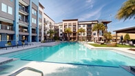 This outdoor amenity space is centered around a generously sized resort-style swimming pool with crystal-clear blue water, serving as the focal point of a well-designed courtyard. The surrounding multi-story building features a contemporary architectural design, combining white and brown exterior finishes with large windows and private balconies that overlook the pool area below.

The pool deck is finished with light-toned pavers and is equipped with multiple lounging areas, including rows of navy blue chaise lounge chairs positioned to maximize sun exposure. Tall palm trees are strategically planted throughout the courtyard, providing a tropical ambiance and natural shade. The shallow entry points and stainless steel handrails offer easy pool access.

Beyond the pool, the space extends into a well-maintained lawn area furnished with additional seating, cabanas with shade canopies, and a covered pergola structure ideal for outdoor gatherings. The courtyard layout creates a sense of enclosure and privacy while still feeling open to the sky. Overall, the space presents a cohesive, upscale outdoor living environment designed for relaxation and recreation.