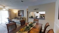This single-story residence showcases an open-concept floor plan that seamlessly connects the dining, living, and kitchen areas into one cohesive living space. The dining area is anchored by a rich, dark wood table with matching chairs, providing an elegant setting for meals and gatherings. A decorative centerpiece of tropical greenery and candles adds a warm, welcoming touch to the space.

A half-wall pass-through with a dark countertop connects the dining area to the kitchen, enhancing the open feel while maintaining a subtle division between spaces. The kitchen features white cabinetry, a classic tile backsplash, and white appliances, contributing to a clean and bright aesthetic. Potted plants along the pass-through counter bring a touch of natural greenery indoors.

The adjacent living room offers a comfortable seating arrangement with a neutral-toned sofa accented by patterned throw pillows. A large framed landscape painting serves as a focal point on the wall, complemented by a traditional floor lamp that adds warm ambient lighting. Textured ceilings and neutral wall tones throughout create a cohesive, understated ambiance suited for relaxed, single-floor living.