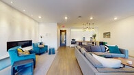 This contemporary single-floor amenity space is thoughtfully designed with an open-plan layout that seamlessly connects multiple functional zones. The main lounge area is anchored by a large, plush sectional sofa and complemented by vibrant teal accent chairs, a mounted flat-screen television, and a sleek media console. A round coffee table and a classic lounge chair add to the room's modern, curated aesthetic.

A standout feature of the space is the fireplace lounge, framed by a floor-to-ceiling textured white surround and flanked by a built-in bench with colorful accent pillows. This intimate seating area provides a warm and inviting retreat within the larger open plan.

The kitchenette, partially open to the main area via a pass-through, is outfitted with stainless steel appliances including an oven, dishwasher, and sink set into a clean white countertop. Warm pendant lights hang above the adjacent dining area, which features a long communal table with bar-height seating, ideal for group gatherings.

Light hardwood flooring runs throughout the space, while recessed lighting and large windows allow natural light to fill the interior. Sheer curtains frame the windows, offering views of the surrounding urban skyline. The overall ambiance is polished, contemporary, and well-suited for both social events and casual gatherings.