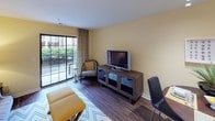 This single-floor residence offers a thoughtfully designed layout with a cohesive warm yellow and gray color palette throughout. The kitchen is a standout feature, boasting rich dark cabinetry, granite countertops, a large island, and a full suite of stainless steel appliances including a refrigerator, dishwasher, microwave, and range — all complemented by hardwood-style flooring and pendant lighting overhead. The open living and dining area flows naturally from the kitchen, featuring the same hardwood-style floors, track lighting over the dining space, and a sliding glass door that fills the room with natural light and provides access to an outdoor area with greenery views. The two bedrooms are carpeted and bathed in soft natural light; the primary bedroom includes windows with blinds and space for a dedicated workspace, while the secondary bedroom offers a serene, relaxed atmosphere. The bathroom is elegantly detailed with granite countertops, a dark wood vanity, a tub-shower combination, and eye-catching patterned wallpaper. A dedicated laundry room with stacked washer and dryer units, built-in wire shelving, and an ironing board station adds exceptional everyday convenience to this well-rounded residence.