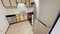 This single-floor space includes a kitchen designed for everyday functionality. The kitchen features a classic layout with wood-trimmed upper and lower cabinetry finished in white, providing ample storage space. Light-toned countertops offer a clean, neutral work surface, complemented by a stainless steel single-basin sink positioned along the lower cabinet run. Appliances include a stainless steel refrigerator, a black and stainless gas range with multiple burners, a dishwasher, and an overhead microwave with a ventilation hood. The flooring consists of neutral-toned stone-look ceramic tiles laid in a grid pattern, lending a durable and easy-to-maintain surface to the space. Under-cabinet lighting above the sink area provides focused task illumination. The overall aesthetic of the kitchen is practical and straightforward, combining classic wood-accented cabinetry with stainless appliances for a timeless, no-frills design suited to daily use.