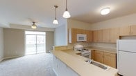 This single-floor space showcases an open-concept layout that seamlessly connects the kitchen and living area. The kitchen is well-appointed with light maple cabinetry offering generous storage, a white gas range, built-in microwave, and a double-basin sink integrated into a spacious wraparound peninsula with a light-toned laminate countertop. The peninsula provides additional prep space and serves as a natural divider between the kitchen and the adjoining living area.

Two pendant lights hang above the peninsula, providing focused task lighting, while a flush-mount ceiling fixture supplements overall illumination. The living area extends comfortably beyond the kitchen, featuring neutral carpeting and a ceiling fan that enhances air circulation. A sliding glass door floods the space with natural light and opens directly onto a balcony, offering a pleasant outdoor extension of the interior.

The overall aesthetic is clean and understated, with light-toned finishes throughout, including soft neutral walls and consistent flooring transitions that contribute to a cohesive, airy ambiance well-suited for everyday living.