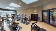 This fitness center is a thoughtfully designed, single-floor facility built to support a full cardio and general fitness routine. The space features light hardwood flooring contrasted by dark rubber equipment mats, creating a clean and functional aesthetic throughout. Neutral warm-toned walls are accented by stylish wall-mounted uplights that complement the recessed ceiling fixtures, contributing to an inviting and energizing atmosphere.

A standout architectural feature is the row of skylights running along the ceiling, which flood the space with natural light and lend an open, airy quality to the room. The generous ceiling height further enhances the sense of spaciousness.

The cardio equipment selection is extensive, with multiple Matrix-brand treadmills, elliptical cross-trainers, and upright and recumbent stationary bikes arranged across the floor in an organized layout. A dedicated stretching and floor exercise zone is equipped with foldable exercise mats, providing space for warm-ups, cool-downs, or bodyweight training.

Additional amenities include a wall-mounted flat-screen display, a modular storage shelving unit, and a small stone accent wall that adds a refined architectural detail. Fitness center rules are prominently posted, underscoring a professional and well-managed environment. The overall ambiance is modern, clean, and functional — ideal for a focused workout experience.