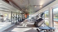 This fitness center is designed to accommodate a comprehensive range of workout needs within a single-story layout. The main exercise area is an elongated, open space with a textured exposed ceiling finished in a dark, industrial tone, complemented by track and pendant lighting that adds warmth throughout the room. Large windows line the perimeter walls, allowing generous natural light to pour in while offering outdoor views of surrounding greenery.

The cardio zone features Matrix-brand stair climbers, ellipticals, and treadmills arranged along the windowed walls. Opposite these machines, a row of power racks with barbells and a full dumbbell rack serves the strength training area, with floor-to-ceiling mirrors amplifying the sense of space. A cable machine station provides additional functional training options.

A bold, curved orange and white mural serves as a striking focal point on the rear accent wall, adding a vibrant, modern aesthetic to the space. Rubber flooring throughout the main area ensures durability and safety during workouts.

Adjacent to the main floor is a separate studio room featuring light hardwood flooring, a ceiling fan, full mirrored wall, and direct access to an outdoor pool area, making it well-suited for yoga, stretching, or group fitness classes.