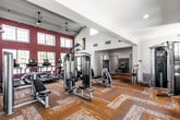 This single-floor fitness center is thoughtfully designed to accommodate a full range of workout needs in a bright, welcoming environment. The main exercise area features soaring ceilings with large, multi-pane windows set against bold crimson accent walls, flooding the space with natural light and offering outdoor views. Ceiling fans provide added comfort throughout. The patterned gold and grey carpet tiles add a modern, dynamic aesthetic to the main floor.

The cardio zone is outfitted with multiple treadmills and Precor elliptical machines, all positioned to face the expansive windows. A dedicated strength training area hosts a Precor cable machine, weight benches, and additional resistance equipment, providing a comprehensive selection for full-body workouts.

A separate functional training studio features durable wood-look flooring, floor-to-ceiling mirrors, and a ceiling fan. This room is equipped with TRX suspension trainers, a full dumbbell and kettlebell rack, a punching bag, a mini trampoline, and a wall-mounted Octagon functional training rig, making it ideal for high-intensity and cross-training workouts.

Additional amenities include a stainless steel water fountain and bottle-filling station set against a stylish penny-tile accent wall, as well as an accessible restroom. The facility also features a bank of secure lockers, completing the full-service gym experience.