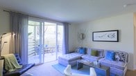 This single-floor space centers around a well-appointed living room designed for both comfort and style. The room is filled with natural light, courtesy of large sliding glass doors fitted with sheer curtains that lead directly to a private outdoor patio. The patio, visible through the glass, features seating and is surrounded by lush greenery, offering a pleasant outdoor extension of the living area.

Inside, the living room is furnished with a plush, tufted sectional sofa accented with decorative blue and green throw pillows, complemented by a matching ottoman and a sleek glass-top coffee table. A blue area rug grounds the seating arrangement, adding a cohesive color palette to the space. The walls are kept neutral, allowing decorative accents — including a striking sunburst mirror and a framed bird print — to serve as focal points.

The carpeted flooring, light-toned walls, and white ceiling contribute to an airy, open ambiance throughout. The overall aesthetic is contemporary and inviting, balancing functional furnishings with tasteful decorative touches that create a cohesive and comfortable living environment.