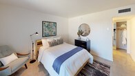 Located at 1116 W Indian School Rd in Phoenix, Arizona, this single-floor unit offers a clean, modern aesthetic with Southwestern-inspired design details. The bedroom serves as the centerpiece of the space, featuring crisp white walls that maximize brightness and a large sliding glass door that floods the room with natural light while providing direct access to the outdoors. Polished concrete-style flooring adds an earthy, understated elegance throughout. The bedroom is thoughtfully appointed with a platform bed framed by a warm wood headboard, layered with neutral and blue-toned textiles that complement the space's relaxed palette. A bold abstract artwork in teal and blue tones anchors one wall, while a round mirror atop a dark dresser adds a decorative focal point. A woven Southwestern-style area rug grounds the room with texture and character. A mirrored sliding closet provides practical storage while visually expanding the space. Adjacent, a glimpse into the kitchenette area reveals stainless steel appliances and clean cabinetry. The overall ambiance is calm, well-lit, and cohesive, blending contemporary minimalism with warm desert-inspired accents suited to the Phoenix setting.