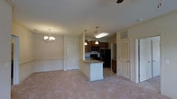 This single-floor residence offers a thoughtfully designed open-concept layout that seamlessly connects the living, dining, and kitchen areas into one cohesive living space. The living and dining area is finished with soft, neutral-toned carpet and features an elegant chandelier that adds a touch of warmth and classic style. A ceiling fan overhead provides practical comfort.

The kitchen is the centerpiece of the space, anchored by a kitchen island with a granite countertop that doubles as a casual dining or prep area. Rich, dark wood cabinetry lines the kitchen walls, complemented by black appliances including a refrigerator, range, and over-the-range microwave. Pendant lights hang above the island, providing focused task lighting and adding visual interest to the space.

The layout includes multiple doorways leading to additional rooms, including what appears to be closet storage with louvered doors, ideal for organization. The neutral beige walls and light-colored ceilings create a bright, airy ambiance throughout. The open flow between areas makes the space feel welcoming and functional, well-suited for both everyday living and entertaining. Overall, the design balances practical functionality with classic residential aesthetic elements.