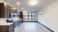 This single-floor unit offers a clean, modern layout designed for functional everyday living. The open-concept main area seamlessly connects the kitchen and living space, creating an efficient and versatile floor plan. Light hardwood-style flooring runs throughout, complemented by crisp white walls and dark navy baseboards that provide a sharp visual contrast.

The kitchen is a standout feature, outfitted with rich dark-toned cabinetry extending from floor to ceiling for ample storage. A full suite of stainless steel appliances includes an electric range, built-in microwave, refrigerator, and dishwasher. Light-colored countertops offer generous prep space, and a single-basin sink with a modern faucet completes the practical setup. Recessed lighting illuminates the kitchen area, while a large window brings in natural light to brighten the adjoining living space.

The living area is open and versatile, with a wall-mounted mini-split climate control unit offering efficient heating and cooling. A large multi-pane window provides natural light and views to the exterior. The entry corridor leads to the main door and connects to the bathroom, maintaining a logical and accessible flow throughout the unit.

The bathroom features a dark wood vanity with a white countertop and integrated sink, a large mirror, and chrome fixtures. Grab bars are installed near the toilet, reflecting an accessible design. Light tile flooring and neutral walls create a clean, understated finish.
