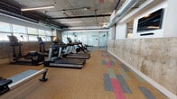 Located at 4065 Glencoe Ave in the Marina del Rey area of Los Angeles, this fitness center showcases a compelling industrial-modern aesthetic. The space features exposed concrete ceilings with visible ductwork and structural columns, lending it a raw, urban character that is complemented by clean white walls and colorful patchwork carpet tile flooring in warm neutrals, reds, and blues.

The main workout floor is generously proportioned, accommodating a robust array of Life Fitness cardio equipment including multiple treadmills, stationary bikes, elliptical trainers, and a rowing machine. A dedicated strength-training section provides weight benches and cable machines, offering a well-rounded fitness experience. Glass-partitioned walls divide areas within the facility, creating distinct zones while maintaining an open, airy feel throughout.

Natural light filters in through a row of windows along one wall, brightening the space and providing exterior views. Multiple wall-mounted flat-screen televisions are positioned throughout for convenience during workouts. Linear LED lighting runs across the ceiling, ensuring consistent illumination. A hydration station with water fountains and a towel service area with decorative tiled accent wall adds a thoughtful, amenity-focused touch. The space is accessible via a stairwell exit and is designed to serve a professional or residential building community.