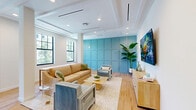 Located at 125 Quadrille Plaza Dr in West Palm Beach, FL, this thoughtfully designed single-floor amenity space blends modern coastal aesthetics with functional versatility. The open layout flows seamlessly across five distinct zones, all unified by warm light hardwood flooring, crisp white ceilings with recessed lighting, and a cohesive palette of soft blues and neutrals.

The main lounge area features a coffered ceiling detail, large black-framed windows that invite abundant natural light and garden views, and a signature teal wainscoted accent wall. Comfortable seating — including a tan sofa, upholstered armchairs, and a central coffee table — surrounds a wall-mounted flat-screen display, creating an inviting social hub.

Adjacent to the lounge, a banquette dining area offers continuous bench seating along another paneled blue accent wall, accented by warm gold wall sconces. Round marble-topped bistro tables paired with woven rattan chairs lend a refined yet relaxed atmosphere to this gathering spot.

The kitchen is well-appointed with light wood and gray cabinetry, a large quartz-topped island, stainless steel appliances, an undermount sink, open shelving, and a striking blue botanical tile backsplash. French doors at the far end provide direct access to an outdoor terrace, filling the kitchen with natural light and greenery views.