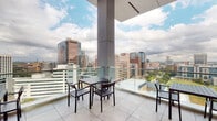 This elevated amenity space combines a generous open-air terrace with a sophisticated interior lounge, offering residents a well-appointed environment high above the surrounding urban landscape. The outdoor terrace is finished with large-format porcelain tiles and enclosed by sleek glass railings that preserve unobstructed panoramic views of the city skyline, parks, and surrounding high-rise developments. Multiple seating arrangements, including dark-framed dining tables with chairs and Adirondack-style lounge chairs, are thoughtfully distributed across the terrace to accommodate both social gatherings and relaxed individual use. Structural columns integrated into the design provide partial overhead coverage in select areas, offering shade without compromising the open-air atmosphere.

The interior lounge is equally refined, featuring soaring ceilings clad in large marble-effect tile panels that serve as a dramatic focal point, with a wall-mounted display screen adding a modern, functional element. The floor continues the same large-format tile found on the terrace, creating a cohesive visual flow between indoor and outdoor areas. Floor-to-ceiling glazed walls connect the lounge directly to the terrace, flooding the interior with natural light while maintaining sweeping city views. Contemporary dark and navy-toned furnishings, including lounge chairs, benches, and café-style tables, complete the space with a clean, urban aesthetic.