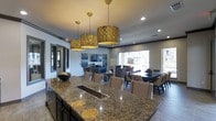 This single-floor space offers an inviting open-concept layout designed for both gathering and entertaining. At its heart is a large, richly patterned granite-topped island complete with an integrated sink, anchoring the space with an air of sophistication. Three oversized ornate gold drum pendant lights hang above, casting a warm glow that complements the recessed ceiling lighting throughout.

The layout flows seamlessly from the island into distinct yet connected zones. A formal dining area features a dark wood table surrounded by upholstered chairs with nailhead trim detailing, while a more casual lounge area provides a relaxed seating arrangement with comfortable armchairs and a wall-mounted television. Large windows at the far end of the space flood the interior with natural light and offer views to an outdoor area beyond.

The overall ambiance is refined yet welcoming, with a neutral gray color palette accented by warm wood tones and gold metallic finishes. Dark cabinetry grounds the space while decorative elements, including abstract wall art and curated accessories, add personality. French-style doors near the entrance provide an elegant transition point, completing a space that balances functional design with upscale aesthetic appeal.