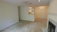 This single-floor residence offers a thoughtfully designed open-concept layout that promotes a sense of spaciousness and easy flow between living areas. The main living space is finished with neutral-toned carpeting that extends throughout, creating a cohesive and comfortable feel underfoot. A ceiling fan is centrally mounted overhead, contributing to both comfort and airflow in the room.

A defining architectural feature is the pass-through kitchen window, which connects the kitchen to the living and dining areas, enhancing the open feel while maintaining a defined cooking space. The kitchen is equipped with white cabinetry and includes standard appliances, with under-cabinet lighting providing functional illumination. Light-colored walls throughout lend a clean, airy atmosphere to the interior.

The dining area is distinguished by an elegant chandelier that adds a touch of classic charm to the otherwise understated design. Recessed lighting supplements the natural and fixture-based illumination across the space. The textured ceiling adds subtle visual interest to the rooms. Overall, the space presents a practical, well-connected single-level layout with a neutral palette well-suited to a variety of furnishing styles and living arrangements.