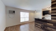 This single-floor space offers a clean, open layout that emphasizes simplicity and functionality. The living area is finished with light hardwood-style flooring that reflects natural light filtering in through a large window dressed with horizontal blinds, lending the room a bright and airy feel. A wall-mounted air conditioning unit provides climate control directly within the living space. Transitioning toward the kitchen area, a standout design element is the rustic bar-style counter clad in reclaimed wood paneling with a dark countertop surface, adding a warm, textured contrast to the otherwise neutral palette of the space. A pendant light hangs above the bar area, providing focused task lighting and a subtle decorative touch. The kitchen is equipped with a range hood, suggesting a dedicated cooking space just beyond the bar counter. The walls are finished in a neutral off-white tone, and the textured ceiling complements the understated aesthetic throughout. Overall, the space presents a practical and welcoming single-level layout with thoughtful design accents, particularly in the kitchen zone, making it a functional and characterful living environment.
