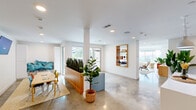 Located in Austin's 78702 zip code, this single-floor community room offers a well-designed, multi-functional gathering space with a modern aesthetic. Polished concrete floors run throughout, reflecting natural light and lending a clean, contemporary feel to the entire area.

The space is organized into two distinct zones. The social and dining area features a wooden dining table with colorful chairs, a built-in planter divider filled with snake plants, a wall-mounted TV, and cozy lounge seating with teal accent chairs. Decorative wall art and open wooden shelving add personality and warmth to the white walls.

The lounge area is a standout, anchored by floor-to-ceiling glass windows that wrap around the exterior, flooding the room with natural light and offering views of the surrounding Austin streetscape and greenery. Curved sofas, modern accent chairs, and patterned area rugs create an inviting, relaxed atmosphere.

The kitchen is fully equipped with dark cabinetry, quartz countertops, a stainless steel refrigerator, and a striking geometric-patterned tile backsplash as a bold focal point. Open wooden shelving adds both function and style. Recessed lighting throughout ensures the space is well-lit at all hours, complementing the abundant natural light from the expansive windows.