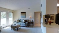 This single-floor space centers around a well-appointed living room that immediately draws attention with its vaulted ceilings, which lend an airy, open quality to the interior. The room is finished in soft, neutral wall tones that complement the plush, light blue upholstered sofa, accented with an assortment of decorative pillows in earthy and floral patterns. A dark wood side table anchors the seating area, topped with a classic table lamp and a small potted plant, adding a touch of warmth and life to the space. An ottoman with a draped throw provides additional casual seating. A framed abstract landscape painting punctuates the wall above the sofa, contributing a subtle artistic element. Plush, light-colored carpeting runs throughout, enhancing the comfortable ambiance. A paneled interior door visible in the background hints at additional rooms beyond. On the opposite side of the living area, a stepped architectural detail suggests a built-in ledge or entertainment area. A wall-mounted speaker is also noted near the ceiling, indicating an integrated audio setup. Overall, the space conveys a relaxed yet refined residential aesthetic.